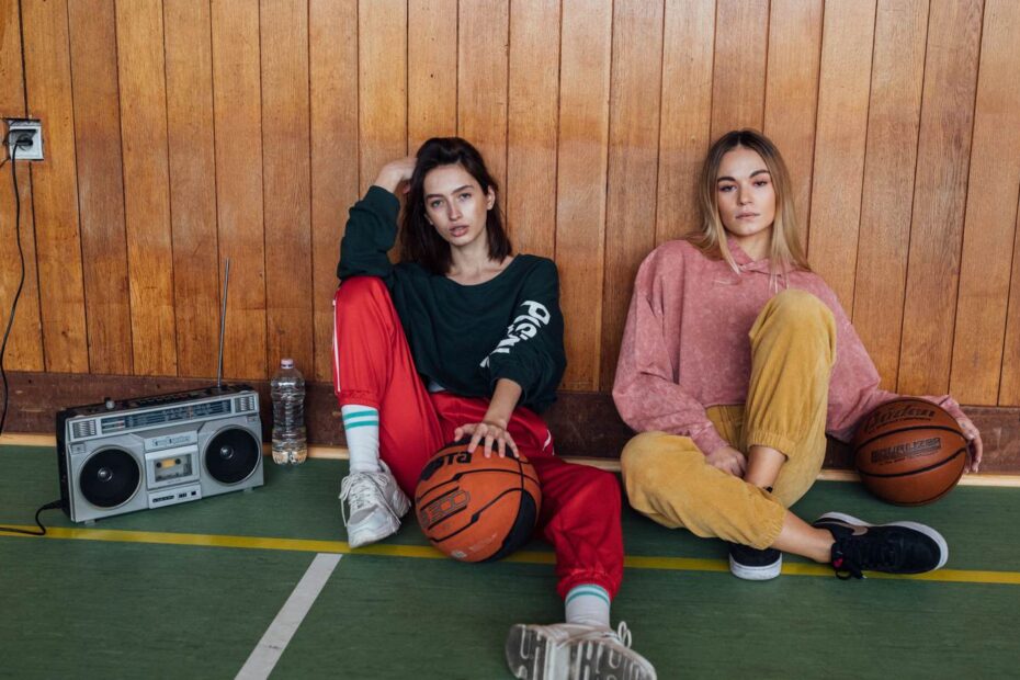02 t2819 Deux femmes en survêtements colorés des années 90 assises devant un mur de bois, radiocassette et ballon de basket