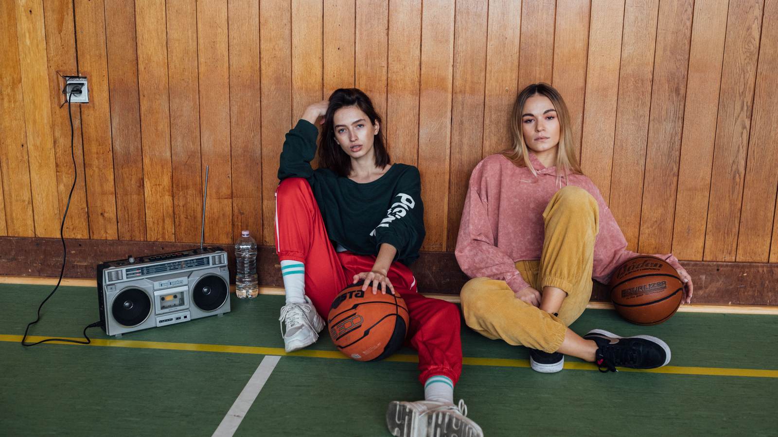 02 t2819 Deux femmes en survêtements colorés des années 90 assises devant un mur de bois, radiocassette et ballon de basket