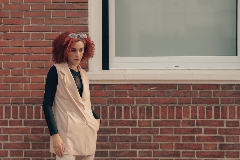 04 t2838 Femme en long manteau sans manches beige porté sur un haut noir, adossée à un mur de brique